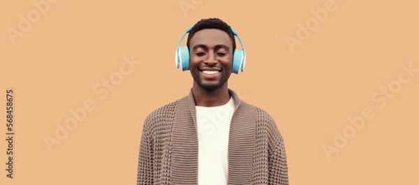 Obraz Portrait of happy smiling african man listening to music in wireless headphones wearing knitted cardigan isolated on brown background