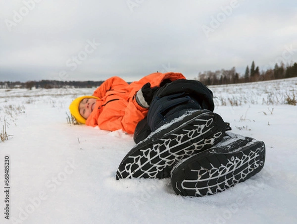 Obraz A child in winter. A boy in a bright orange jacket fell into the snow in a field in nature. The sole is a close-up.