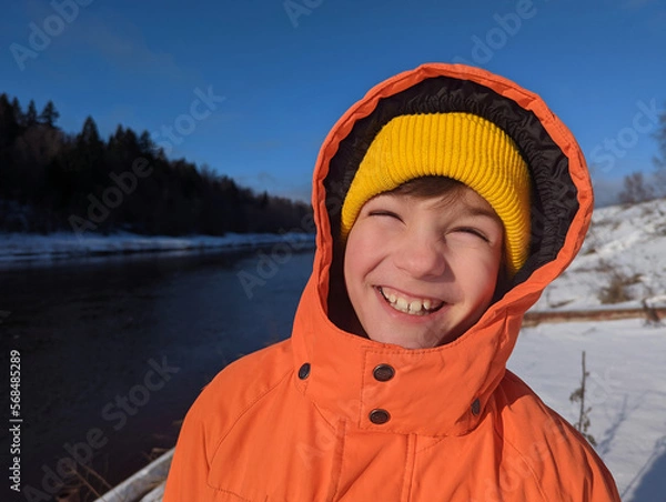 Obraz Portrait of a laughing child in a bright orange jacket against a winter landscape.