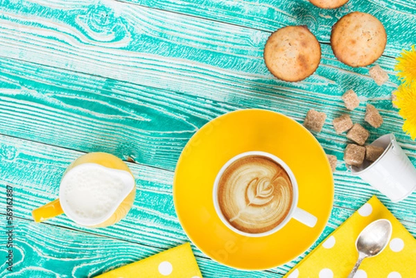 Fototapeta cup of cappuccino coffee on yellow plate and yellow milk jug cane sugar,  cakes, dandelions, teaspoon on turquoise colored wooden table with yellow napkin at polka dots top view
