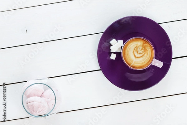 Fototapeta cup of cappuccino coffee on violet plate with sugar,  marshmallows in a glass vase on white colored wooden table , top view