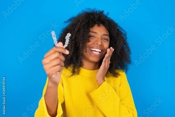Fototapeta Happy young woman with afro hairstyle wearing orange crop top over blue wall holding and showing at camera an invisible aligner while laughing. Dental healthcare and confidence concept.