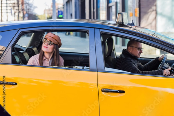Fototapeta woman leaning out of the window of a cab in barcelona, in front of the cab driver driving. pretty girl with cap, sunglasses and pink jacket.