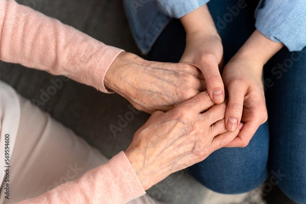 Fototapeta Top view of the mature female aged wrinkled skin and hands of her care giver. Senior woman in elderly care facility gets help from hospital personnel nurse. Grand mother everyday life concept
