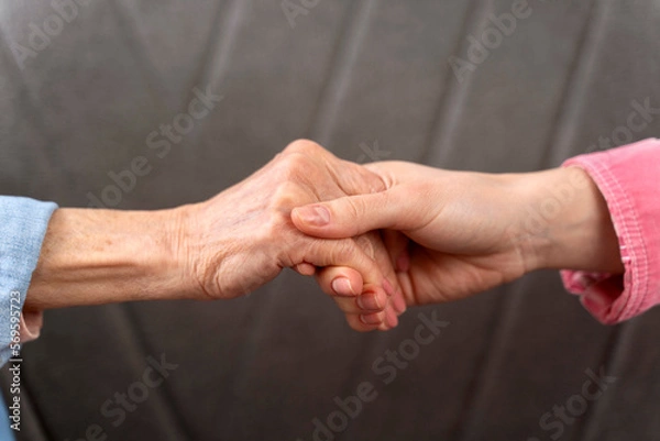 Fototapeta Cropped view of the hands of old woman and her daughter at the sofa background. Family relationships concept