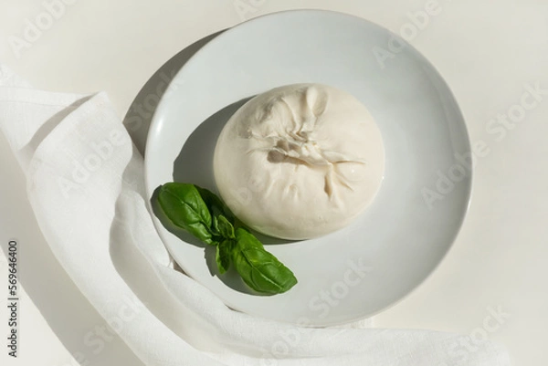 Fototapeta Traditional Italian, fresh, soft burrato ball of young mozzarella cheese with cream on a round white plate with basil greens on a light background. close-up