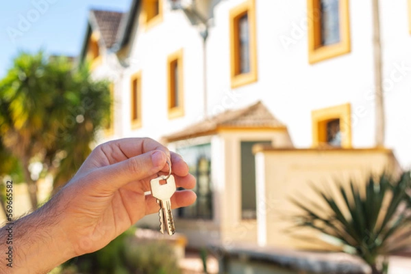 Obraz A man's hand holds the keys to the apartment against the background of the house and palm trees. The topic of buying expensive real estate in countries with a warm climate.
