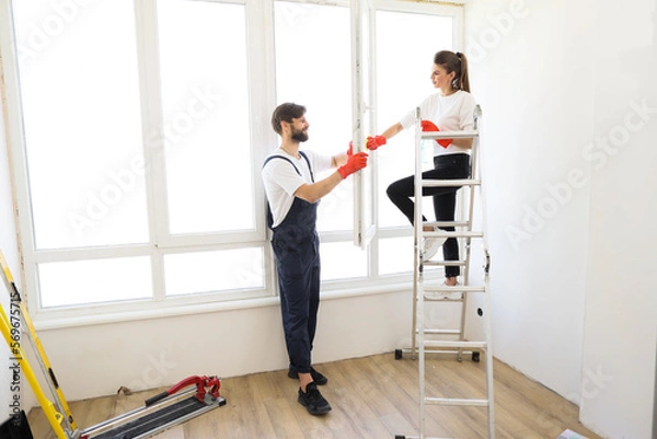 Fototapeta Attractive young couple in rubber gloves with detergent and rag washing windows together. Beautiful woman in casual clothes and bearded man in overalls cleaning their house after repair.