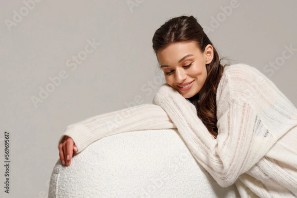 Fototapeta Happy young woman sitting on sofa at home and looking at camera. Portrait of comfortable woman in winter clothes relaxing on armchair. Portrait of beautiful girl smiling and relaxing during autumn.