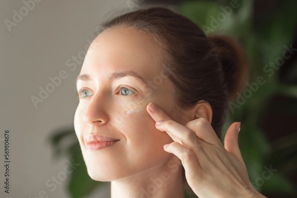 Fototapeta Portrait of beautiful young woman smiling and applying moisturizing face cream or lotion with finger. Skin care concept.