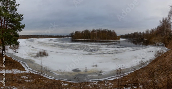Fototapeta Winter. Overcast and cloudy. A sharp bend in the river, ice, gullies in the ice. Islet with a birch grove. Panoramic photography