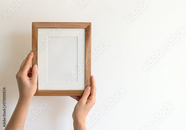 Fototapeta Woman's hands holding a wooden stylish blank frame against a white wall. Frame mock up, copy space for your text.	
