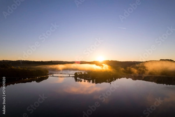 Fototapeta Aerial view of Lac de Saint Pardoux and its footbridge at sunrise with bright yellow cloud