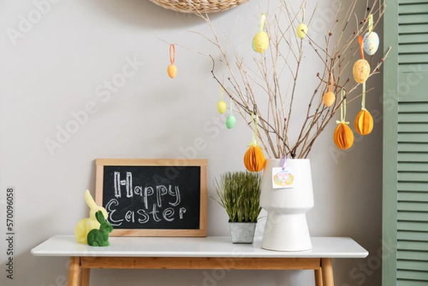 Fototapeta Chalkboard with text HAPPY EASTER, rabbits, grass and tree branches in vase on table near light wall