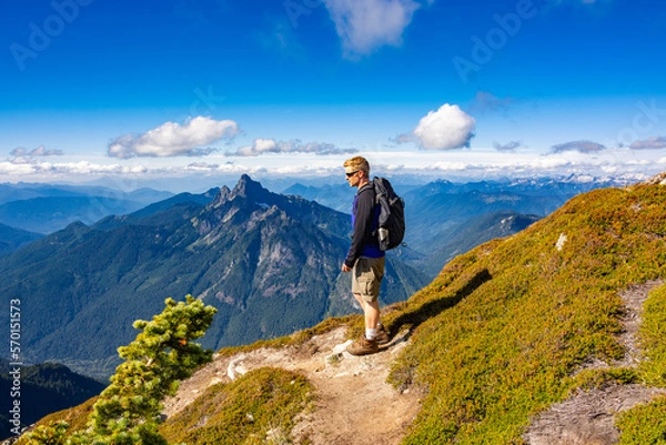 Fototapeta Adventurous athletic male hiker standing on a hiking trail on top of a rugged mountain in the Pacific Northwest with jagged mountains in the background.
