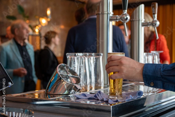 Fototapeta The hand of a caucasian bartender serving a glass of beer from behind his beer tap with people in the background