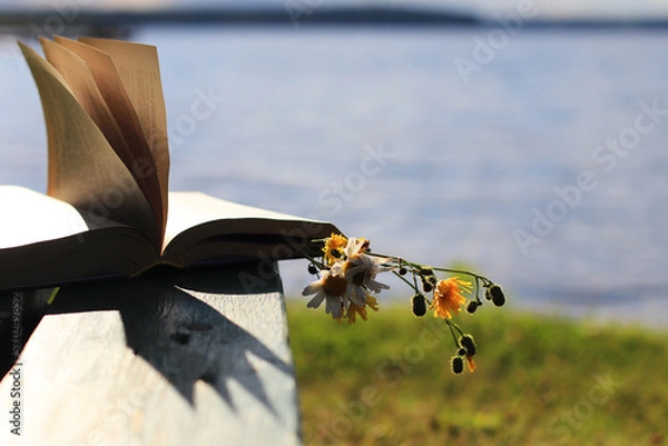 Fototapeta Beautiful bright summer natural background. A bouquet of wildflowers between the pages lies on a bench in the rays of the setting sun. recreation on the banks of rivers