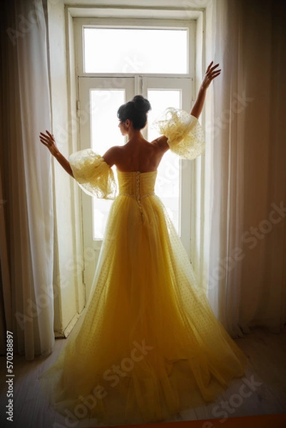 Fototapeta A woman's silhouette in a golden luxurious dress against the background of a window holds a curtain with her hands. Elegant lady in a yellow long silk dress with bare back, back view.