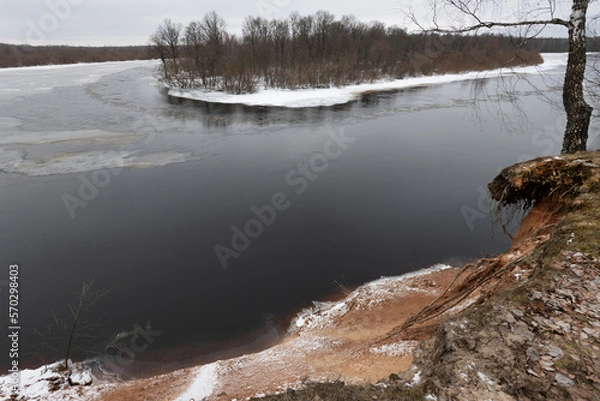 Fototapeta Winter landscape with a partially frozen river. the flow of water. Islet and birch grove. Steep steep bank. Panorama