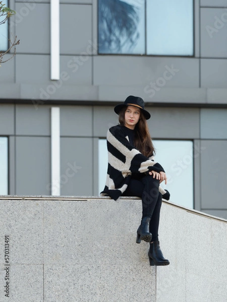 Obraz Stylish young girl with hat is sitting on overhanging wall with legs crossed. Modern building in background. Portrait.