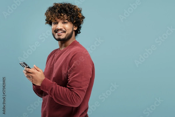 Fototapeta Young  smiling Indian man holding smartphone using mobile app shopping online isolated on blue background, copy space. Portrait of happy asian hipster with curly hair ordering food on website