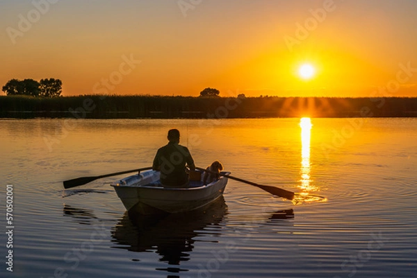 Obraz Silhouette of a 60-year-old Caucasian man with a poodle dog in a traditional rowing boat with oars in his hands against the background of a sunset.