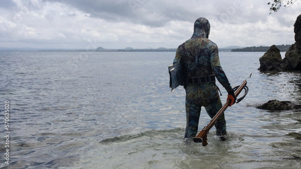 Fototapeta a man in a wetsuit, with camouflage coloring, is preparing for spearfishing, with an underwater rifle in his hands