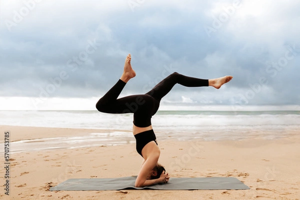 Fototapeta Young woman practicing yoga on the beach by seaside, doing headstand exercise on fitness mat, side view, copy space