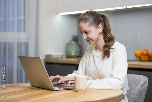 Fototapeta beautiful girl works at home with a laptop at the table and smiles