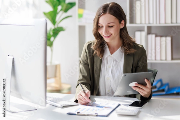 Fototapeta Concept of business working, Businesswoman sitting while writing and checking business analytic document  report  and holding tablet with computer pc, smartphone on desk in her workstation.