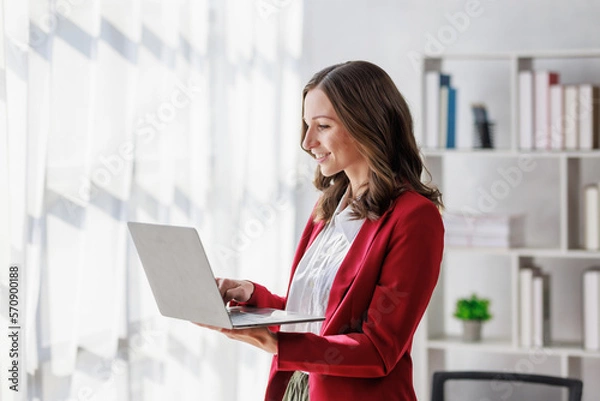 Fototapeta Concept of business working, Businesswoman wearing red suite standing while using computer laptop for checking business analytic document  report  with coffee cup on desk in her workstation.