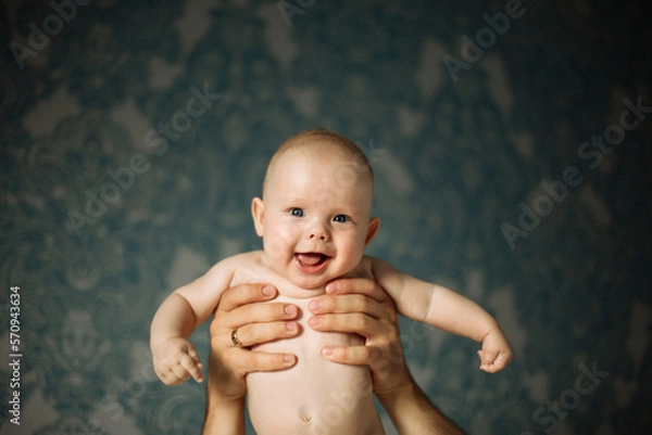 Fototapeta Portrait of happy little baby boy, looking at the camera while hands of the loving parent holding him on background on the wall