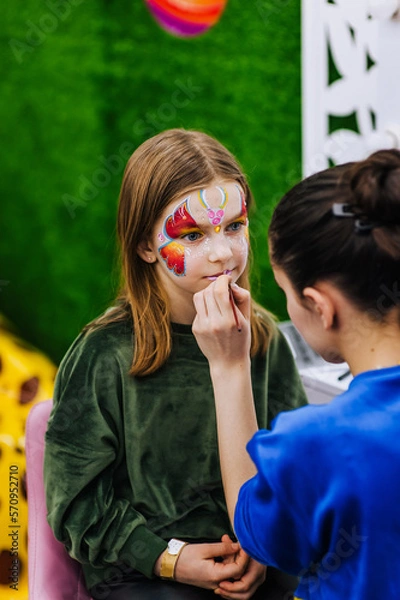 Fototapeta A professional make-up artist, artist paints with a brush on her face with face paints, drawing, children's makeup for a little girl model, a child. Photography, art, concept, lifestyle.