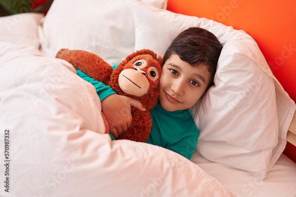 Fototapeta Adorable hispanic boy hugging monkey doll lying on bed at bedroom