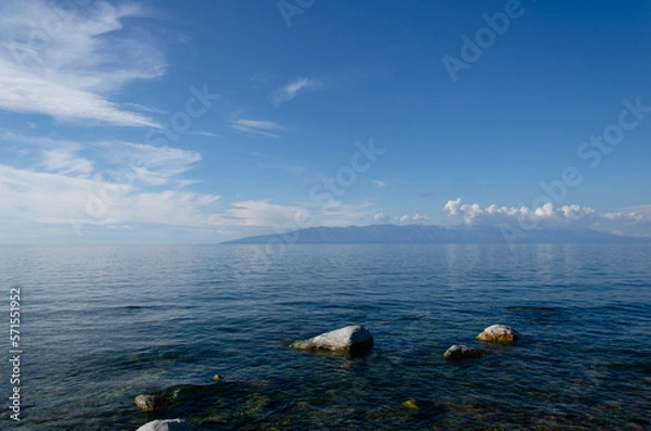 Fototapeta The smooth surface of the water of Lake Baikal on a sunny summer day. Large stones in the water near the shore.