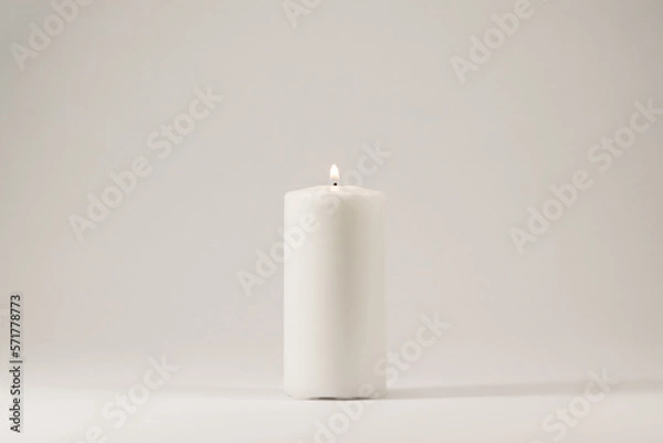 Fototapeta A lighted white candle burning isolated on white background with low light shadow.