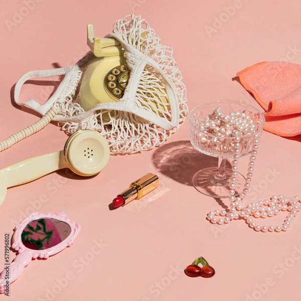 Obraz Rotary dial phone in a mesh bag, lipstick, retro style hand mirror, pearls, coupe champagne glass, creative aesthetic layout, pastel pink background. Nostalgia trend. 