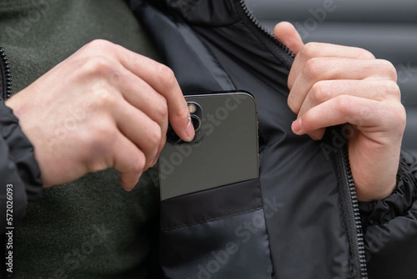 Fototapeta The man puts the phone in the inner pocket of his black jacket. Hands with phone close-up