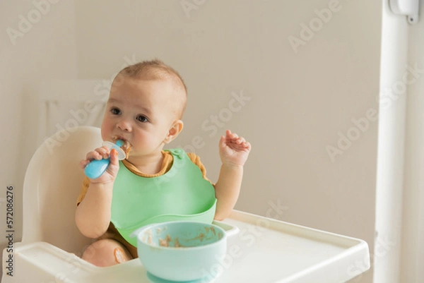 Fototapeta baby 1 year old eats baby food from a spoon sitting at the table, the first complementary foods and healthy food for small children.