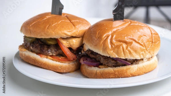 Fototapeta Two homemade hamburgers on white plate, pierced with a dinner knife. The buttery glazed buns and different condiments are in the main focus. Home cooking and burger prep concept.