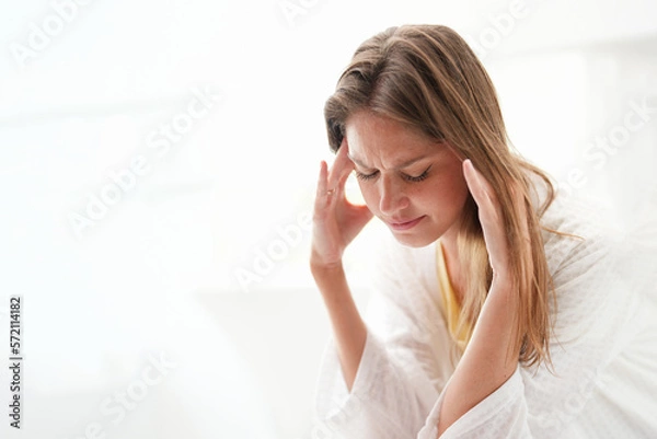Fototapeta Young Caucasian woman is unwell and appears to be experiencing a migraine headache. She is touching  her head while in a brightly lit room. It appears that she is suffering from a health problem.