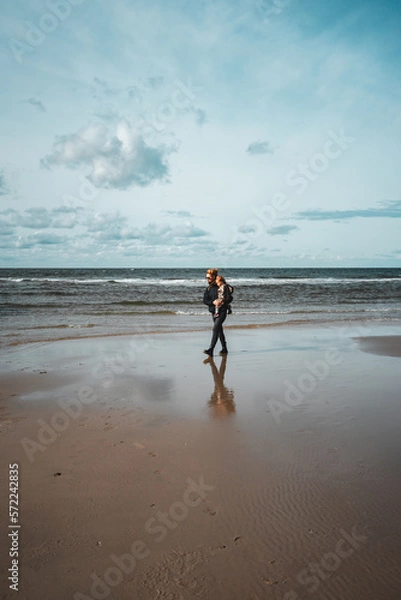 Fototapeta Frau mit Kleinkind auf dem Arm geht am Meer bei Ebbe spazieren, einige Wolken am Himmel