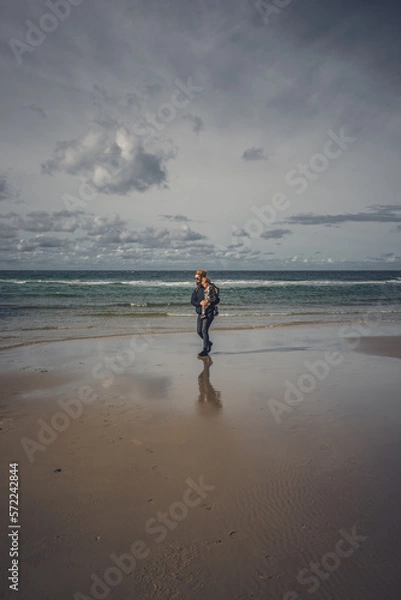 Fototapeta Frau mit Kleinkind auf dem Arm geht am Meer bei Ebbe spazieren, einige Wolken am Himmel