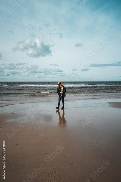 Fototapeta Frau mit Kleinkind auf dem Arm geht am Meer bei Ebbe spazieren, einige Wolken am Himmel