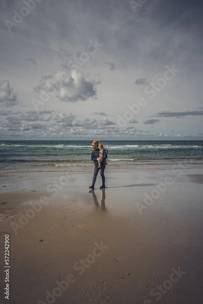 Fototapeta Frau mit Kleinkind auf dem Arm geht am Meer bei Ebbe spazieren, einige Wolken am Himmel