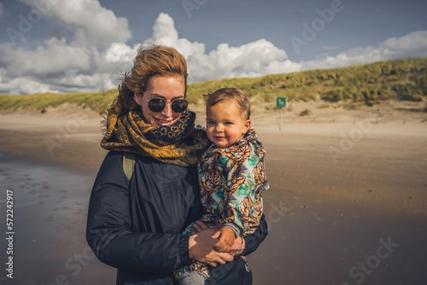 Fototapeta Mutter mit Kleinkind auf dem Arm ab einem Strand an der Ostsee, Wetter ist frisch und der Wind geht stark, trotzdem sind beide fröhlich und lachen 