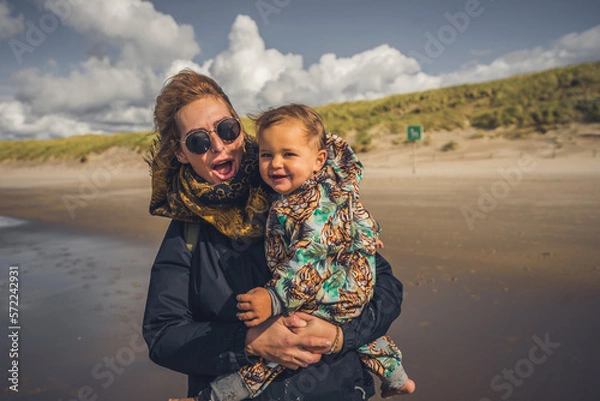 Fototapeta Mutter mit Kleinkind auf dem Arm ab einem Strand an der Ostsee, Wetter ist frisch und der Wind geht stark, trotzdem sind beide fröhlich und lachen 