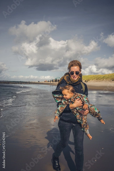 Fototapeta Mutter mit Kleinkind auf dem Arm ab einem Strand an der Ostsee, Wetter ist frisch und der Wind geht stark, trotzdem sind beide fröhlich und lachen 