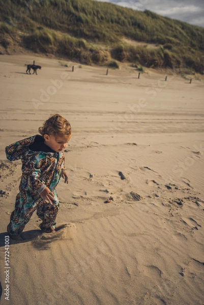 Fototapeta Kleinkind läuft am Stand durch den Sand und hat Spaß, im Hintergrund beobachtet Mutter mit Hund das Mädchen im bunten Overall