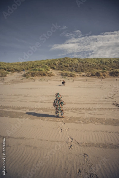 Fototapeta Kleinkind mit buntem Overall läuft am Strand zu seiner Mutter, die am Rand der Dünen auf das Kind wartet